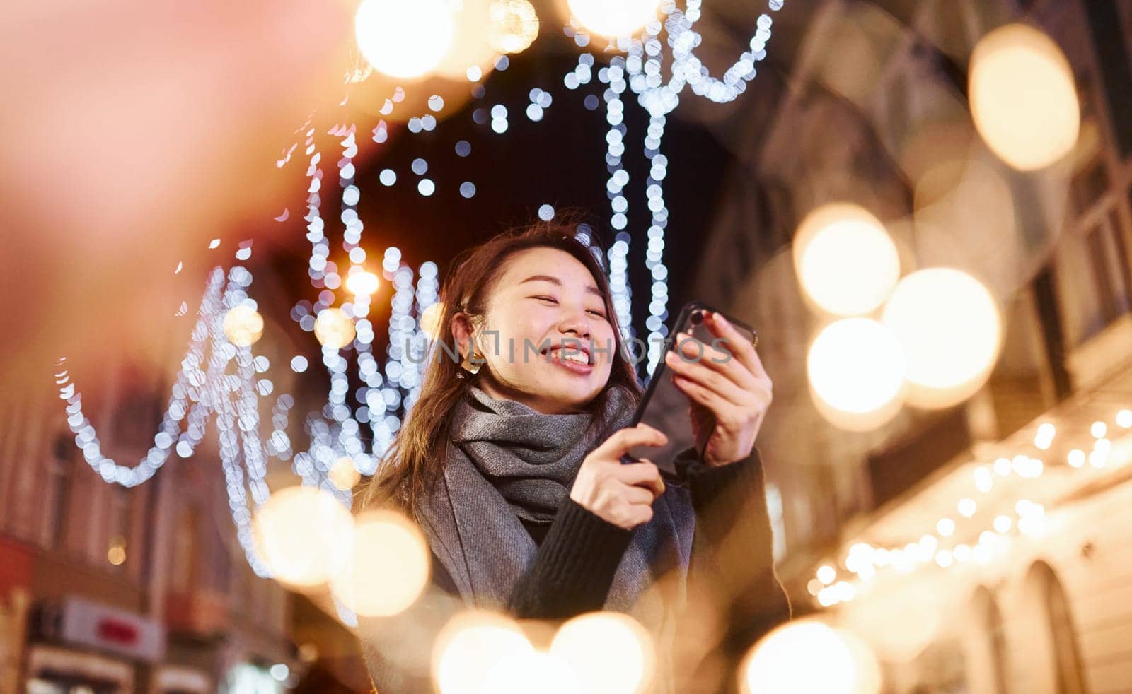 Uses phone. Cute and happy asian young woman outdoors in the city celebrates New year.