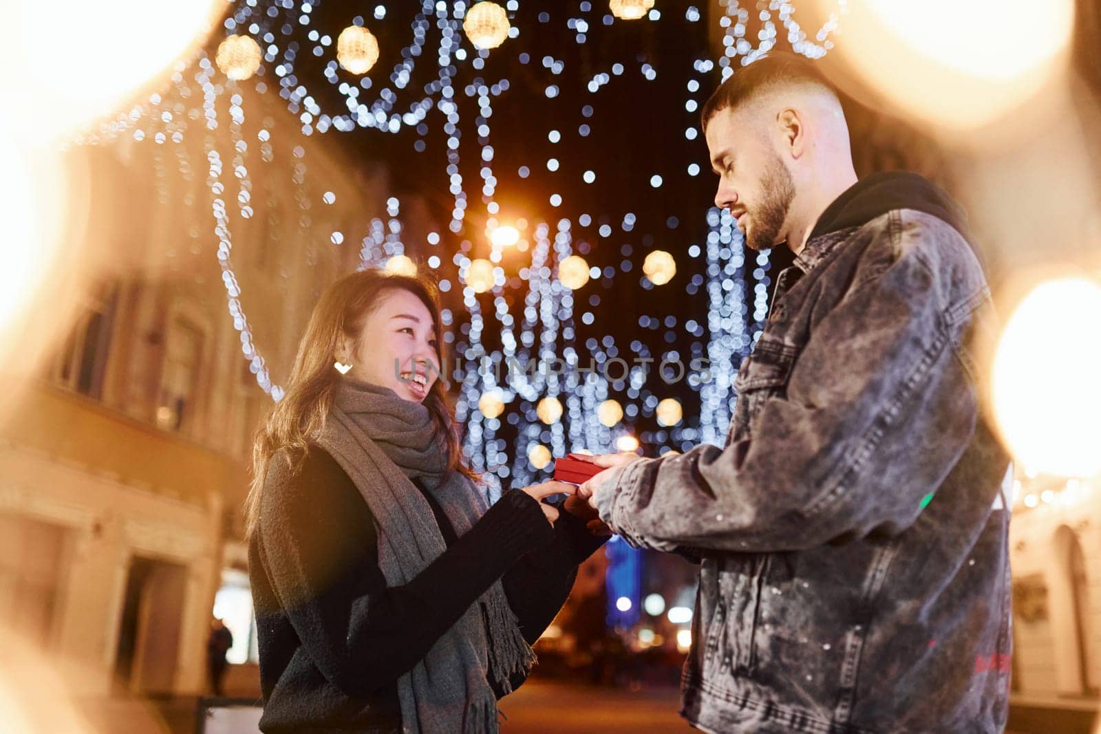 Holds gift box. Happy multiracial couple together outdoors in the city celebrating New year.