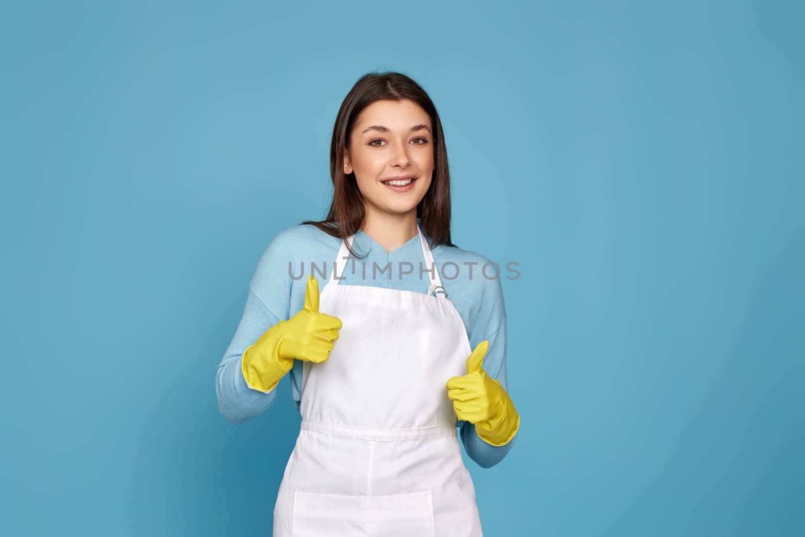 beautiful brunette woman in yellow rubber gloves and cleaner apron showing ok sign on blue background.