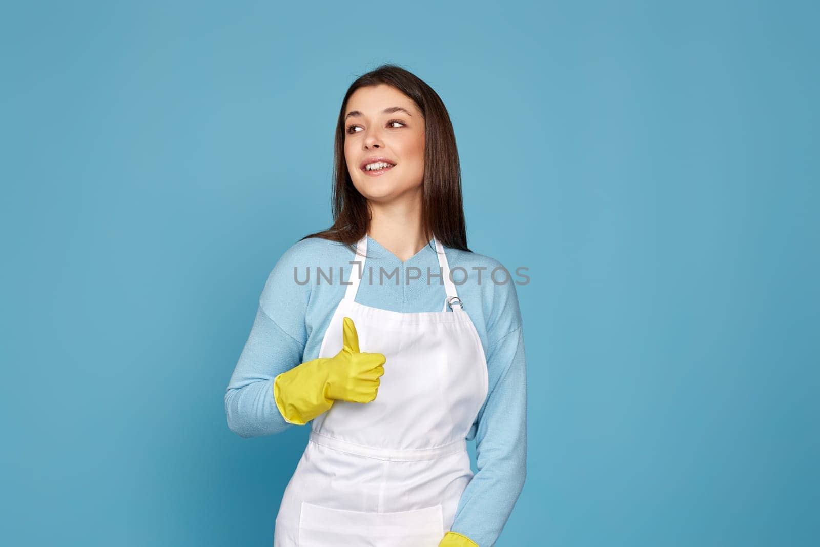 beautiful brunette woman in yellow rubber gloves and cleaner apron showing ok sign on blue background.