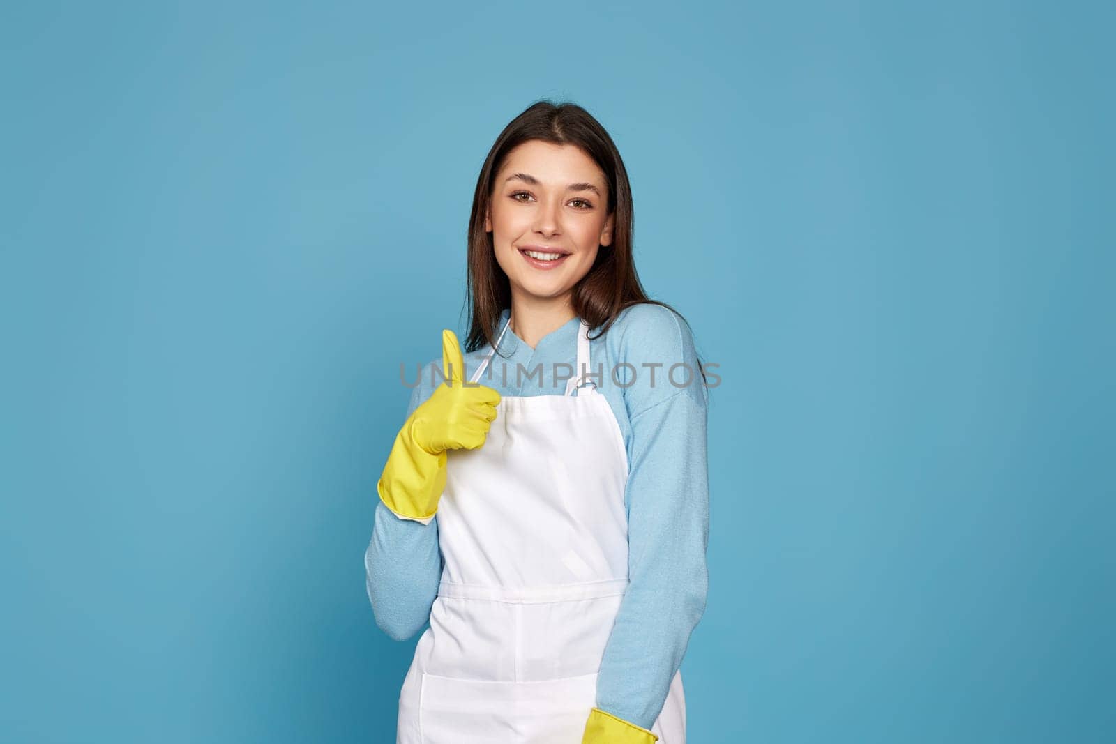 beautiful brunette woman in yellow rubber gloves and cleaner apron showing ok sign on blue background.