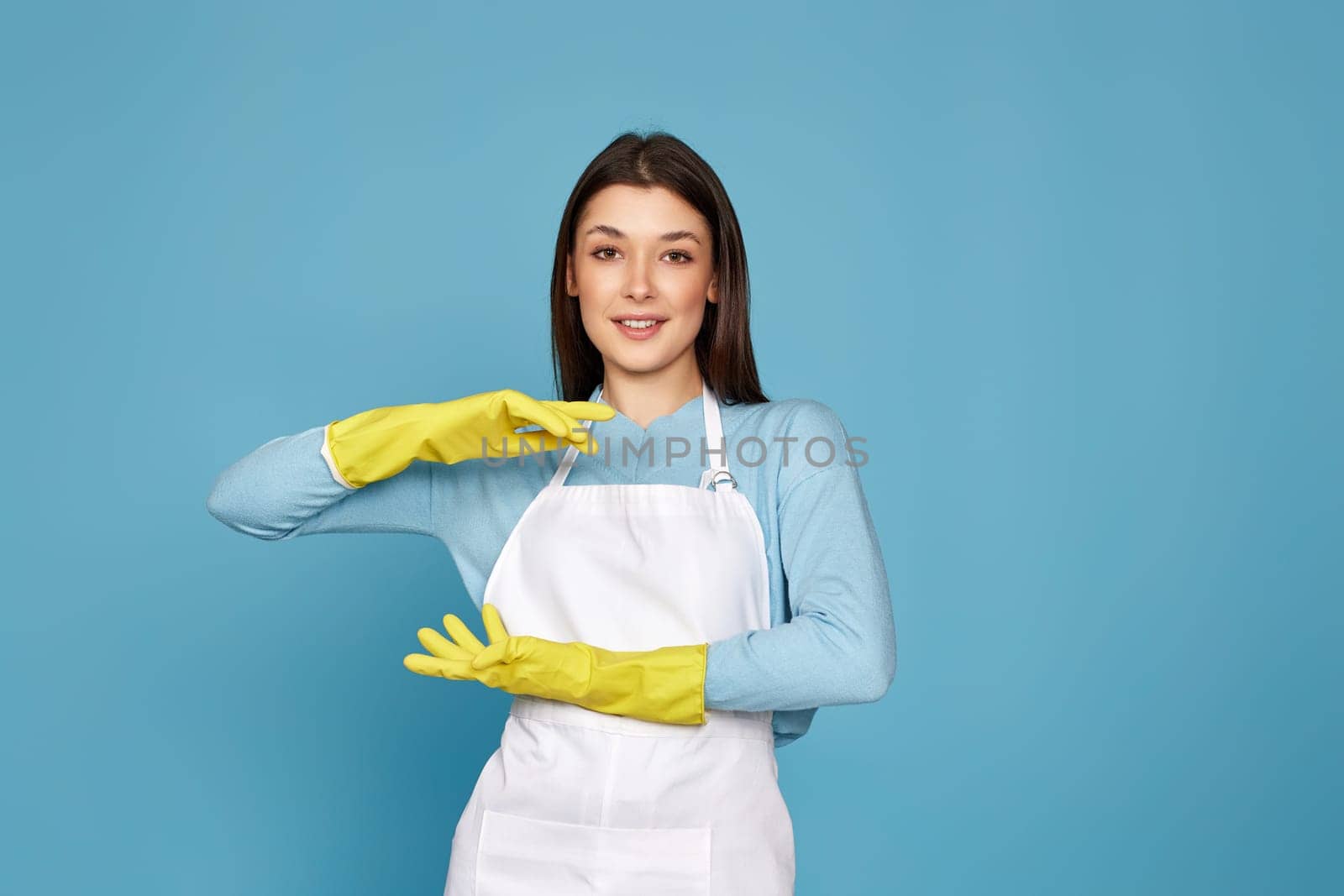 beautiful brunette woman in yellow rubber gloves and cleaner apron showing measure symbol gesture on blue background. cleaning