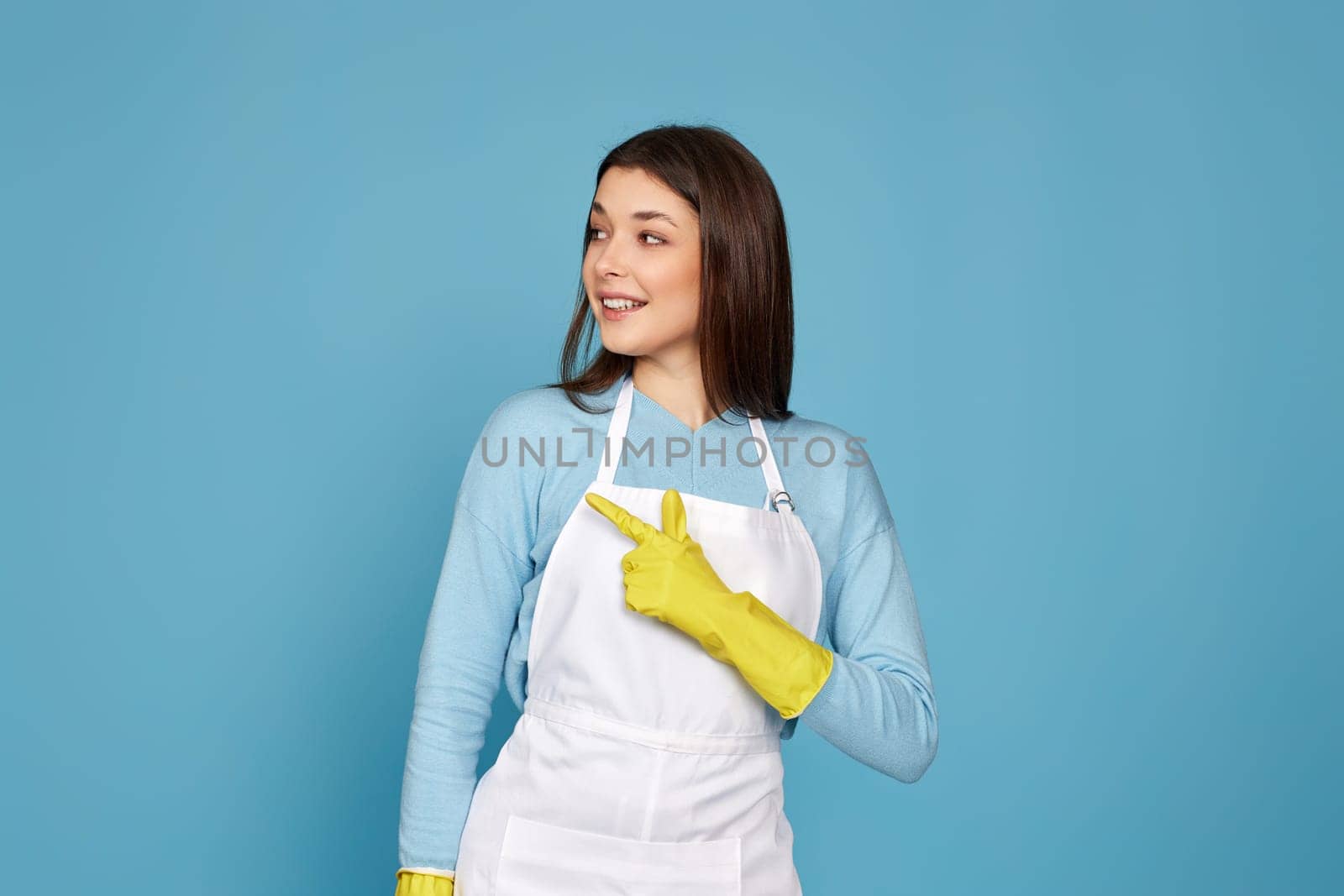 brunette caucasian woman in yellow rubber gloves pointing fingers aside at copyspace isolated on blue background.