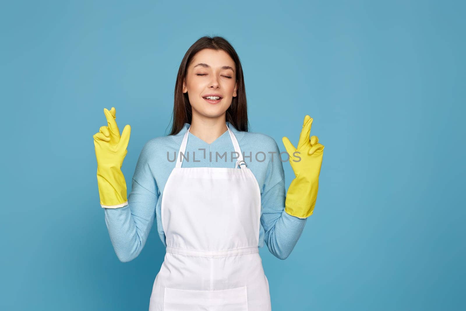 beautiful brunette caucasian girl in yellow rubber gloves with finger crossed gesture isolated on blue background.