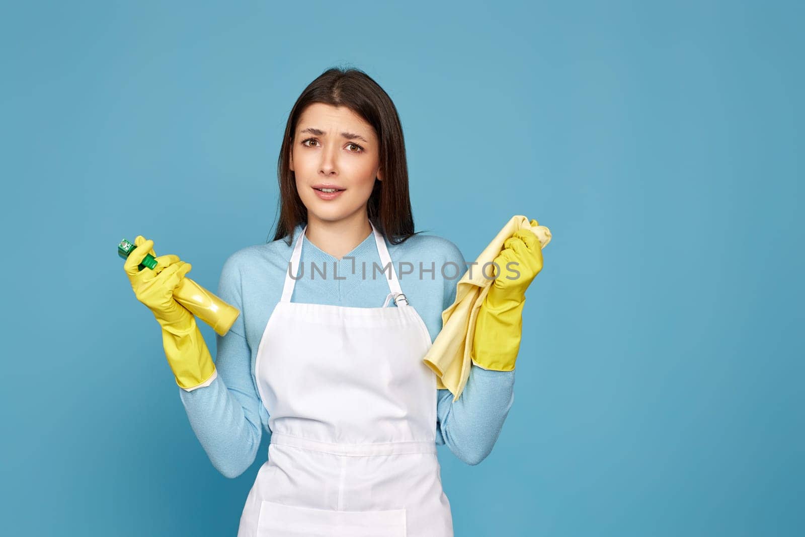 skeptic and nervous, frowning woman in gloves and cleaner apron with cleaning rag and detergent sprayer on blue background.