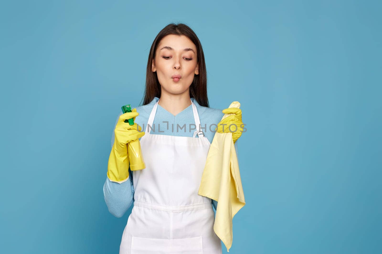 caucasian woman in rubber gloves and cleaner apron with cleaning rag and detergent sprayer on blue background.