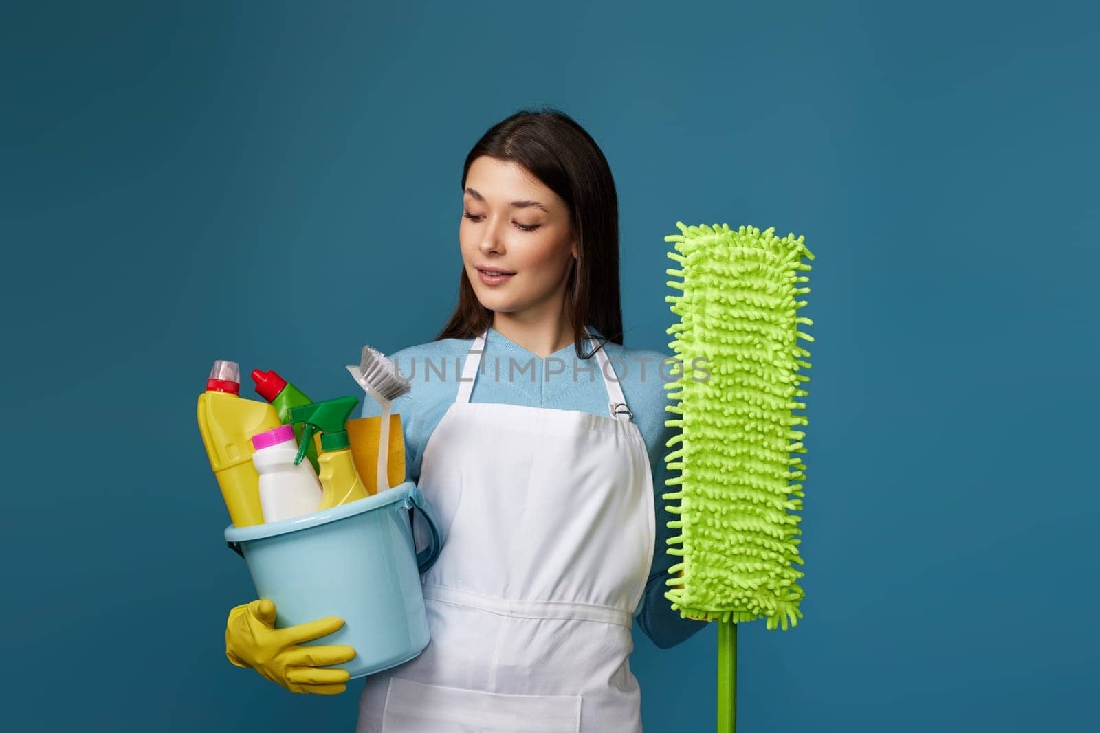 pretty young woman in yellow rubber gloves and cleaner apron holding bucket of detergents and mop on blue background.