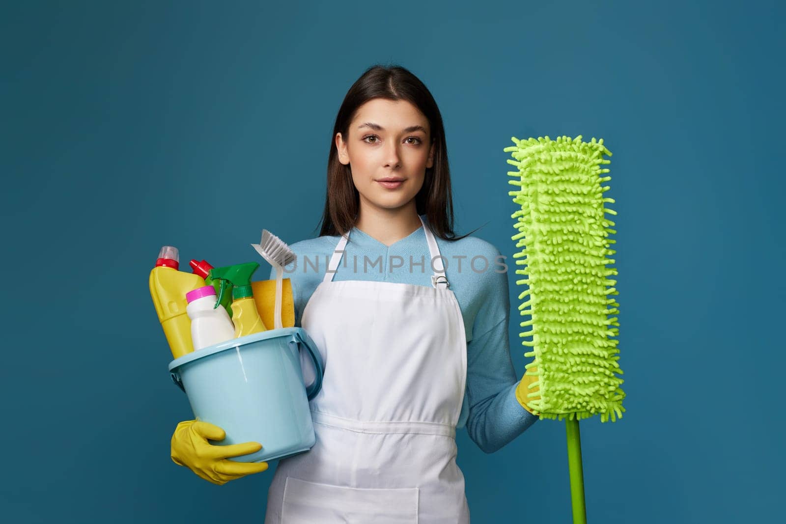 pretty young woman in yellow rubber gloves and cleaner apron holding bucket of detergents and mop on blue background.