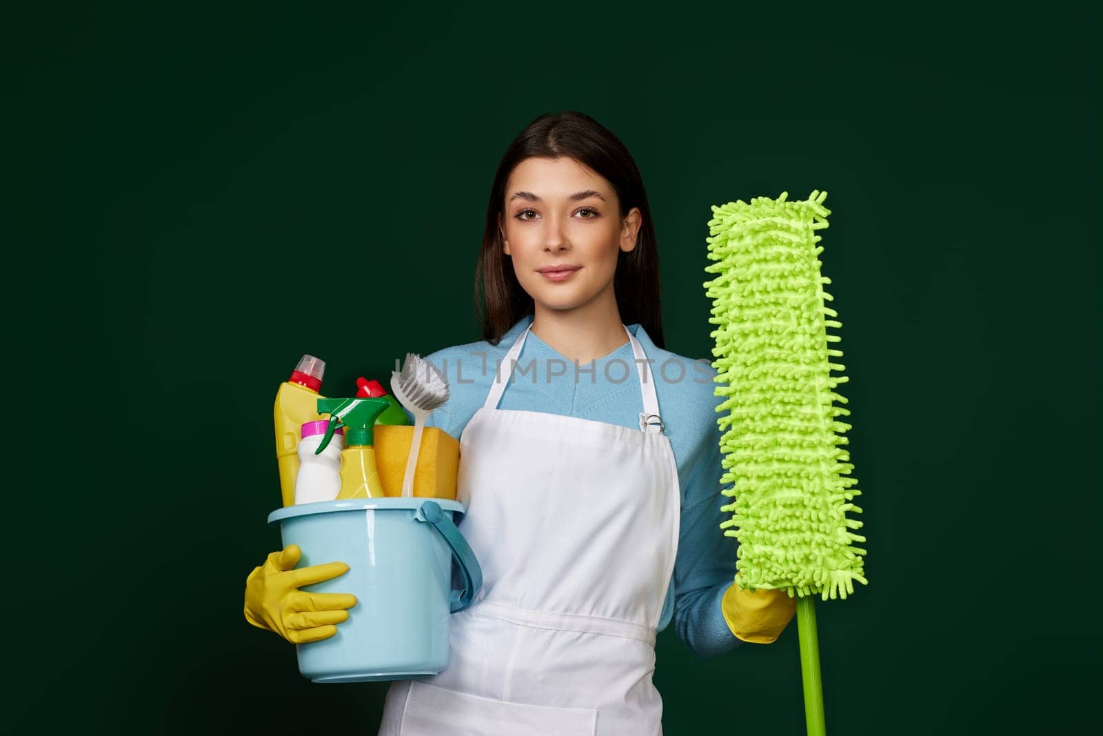 pretty woman in yellow rubber gloves and cleaner apron holding bucket of detergents and mop on dark green background.