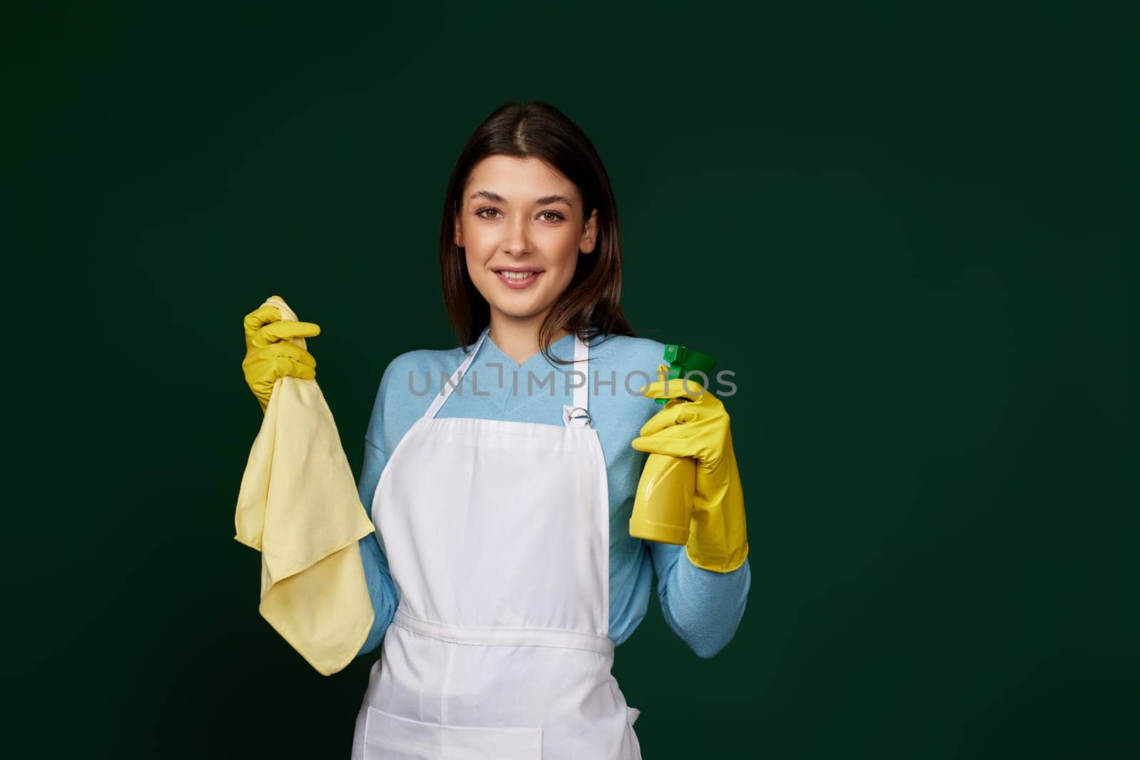happy woman in yellow rubber gloves and cleaner apron with cleaning rag and detergent sprayer on dark green background.