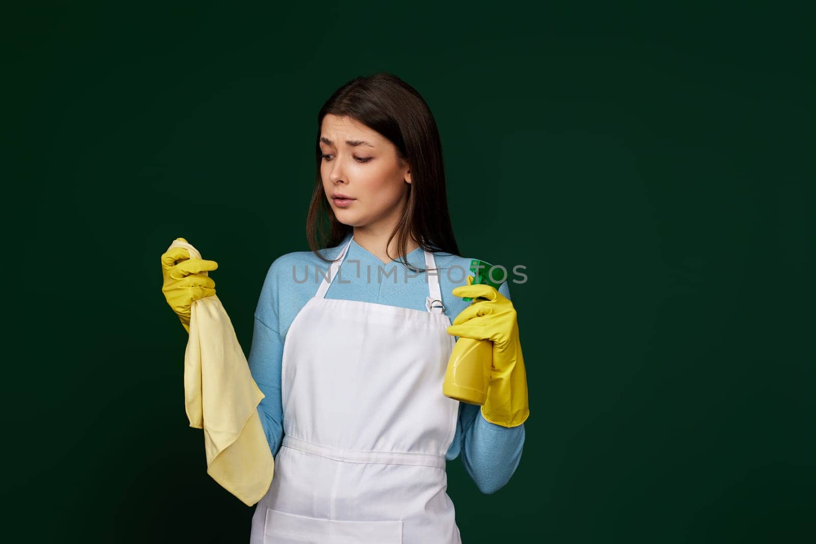 skeptic and nervous, frowning woman in gloves and cleaner apron with cleaning rag and detergent sprayer on green background.