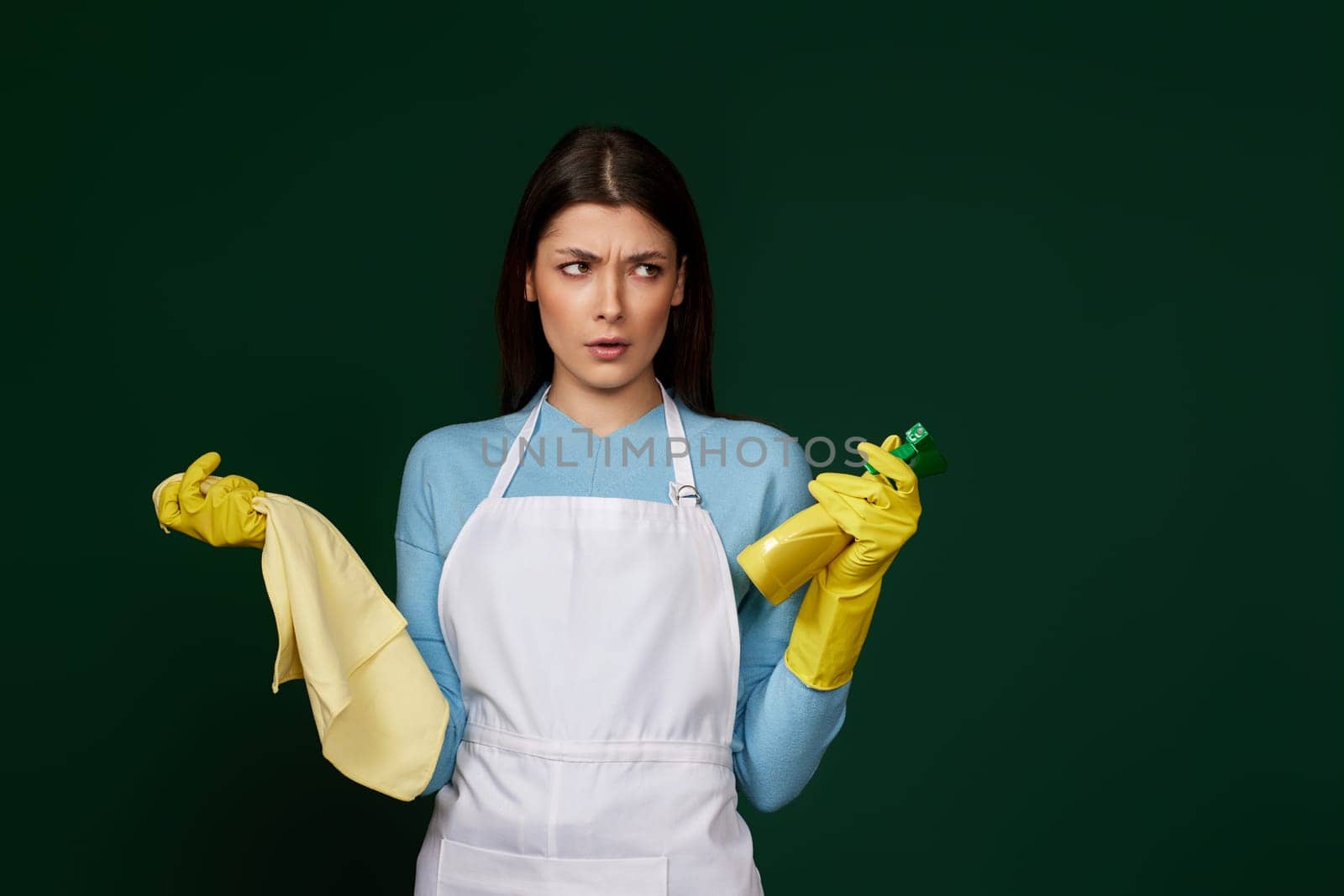 angry woman in gloves and cleaner apron with cleaning rag and detergent sprayer on green background.