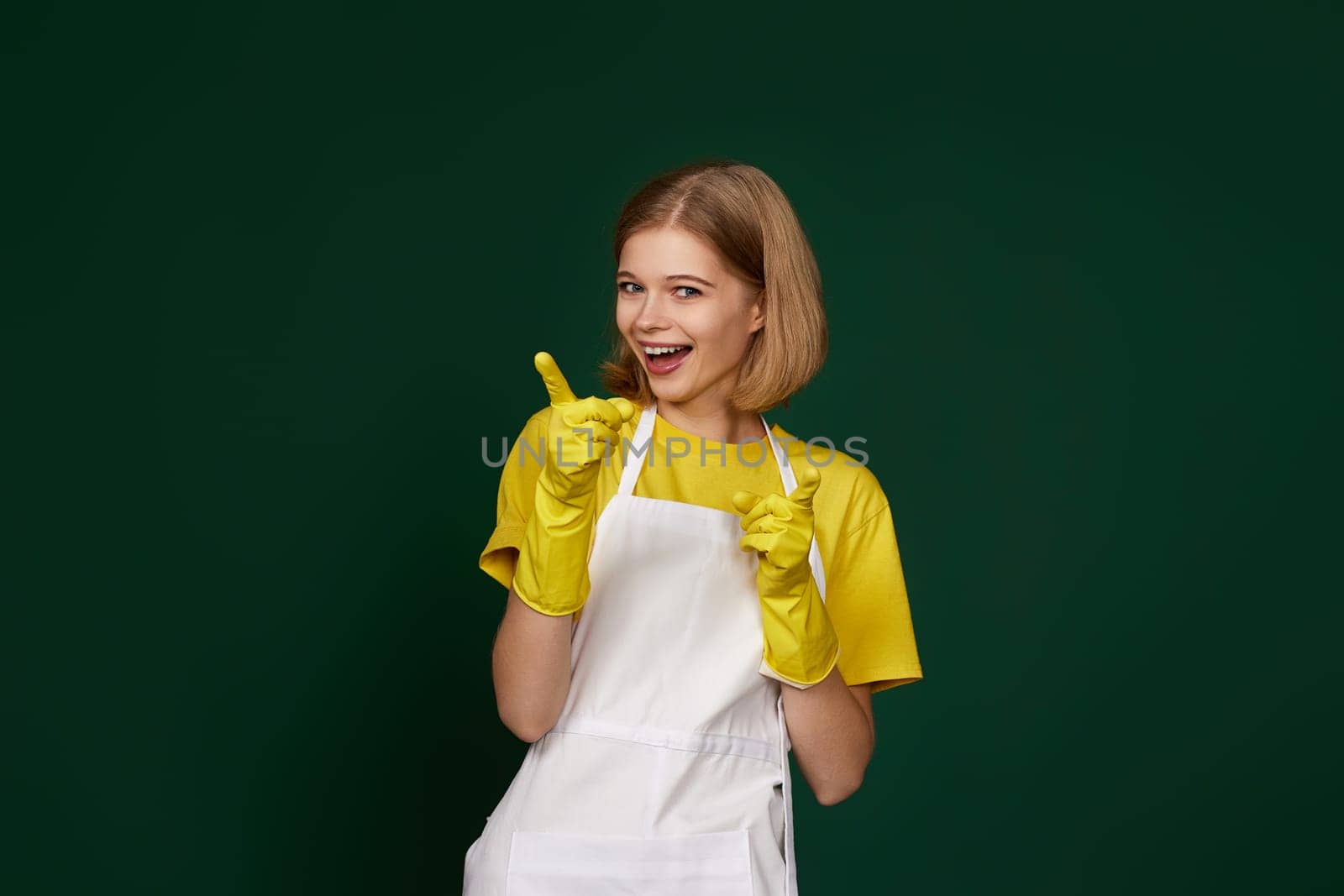 happy blonde woman in yellow rubber gloves and cleaner apron pointing fingers to camera on green background.