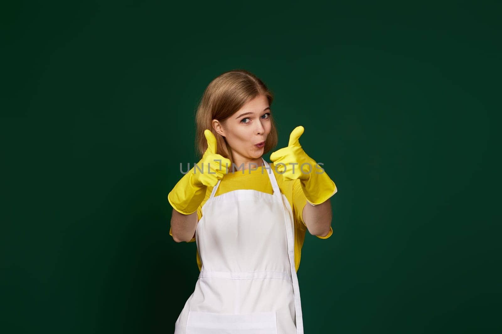 very excited blonde woman in yellow rubber gloves and cleaner apron showing ok sign on green background.