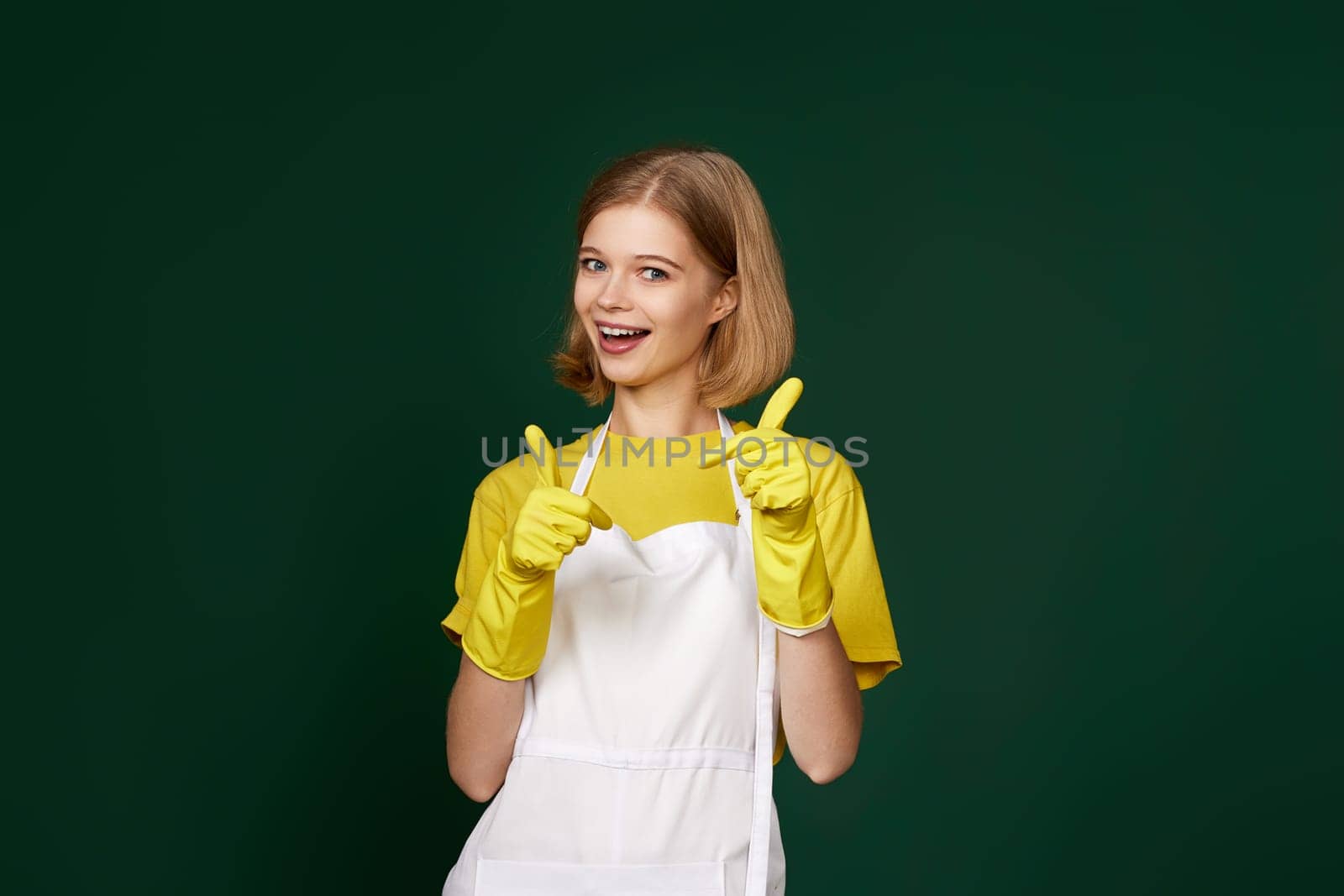 beautiful blonde woman in yellow rubber gloves and cleaner apron showing ok sign on green background.