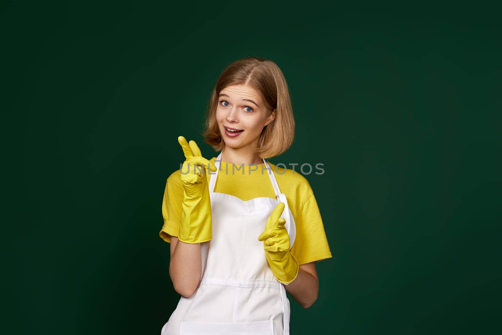 attractive caucasian blonde woman in yellow rubber gloves and cleaner apron pointing fingers to camera on green background.