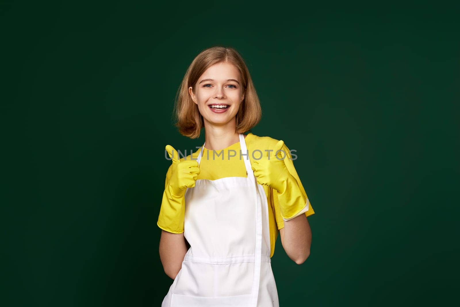 beautiful blonde woman in yellow rubber gloves and cleaner apron showing ok sign on green background.