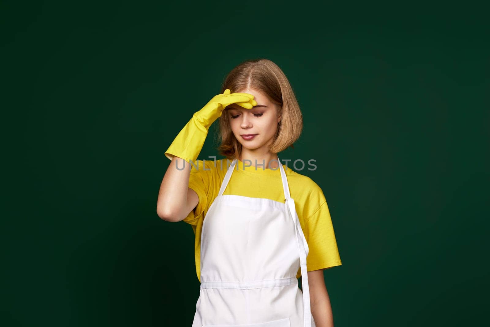 tired blonde woman in yellow gloves and cleaner apron on green background