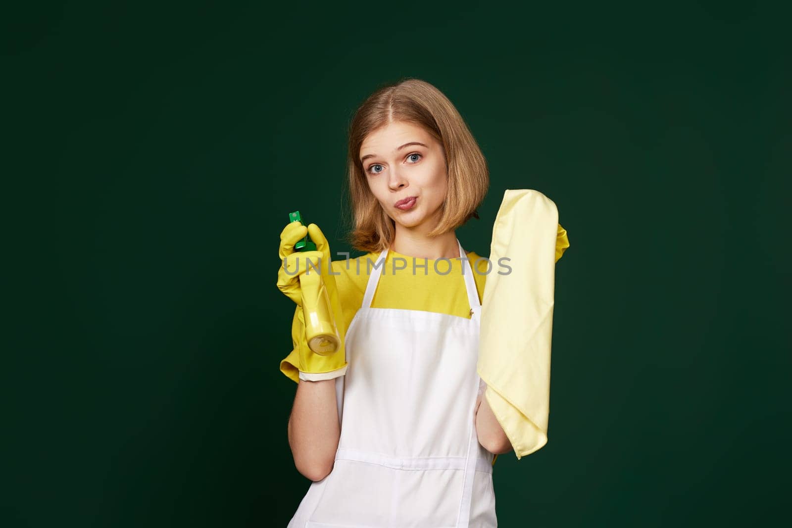 skeptic and nervous, frowning blonde woman in gloves and cleaner apron with cleaning rag and detergent sprayer on green background.