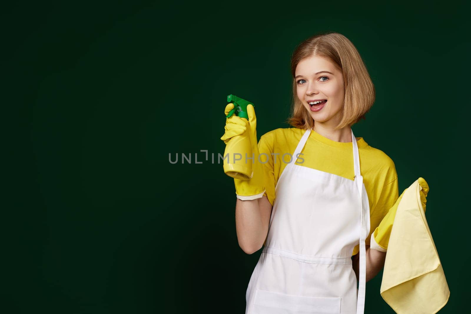 happy blonde woman in yellow rubber gloves and cleaner apron with cleaning rag and detergent sprayer on dark green background.