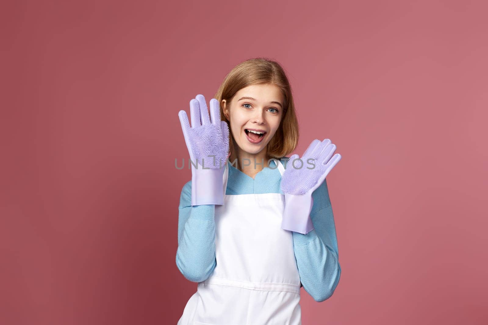 beautiful funny blonde woman in purple rubber gloves and cleaner apron on pink background.