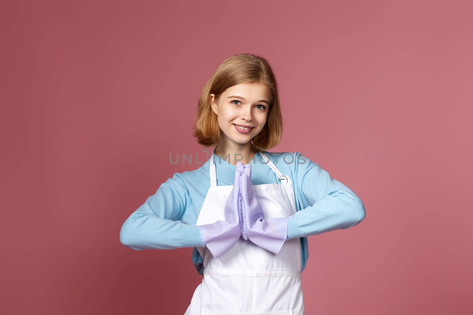 positive blonde woman in purple rubber gloves and cleaner apron praying with hands together on pink background.