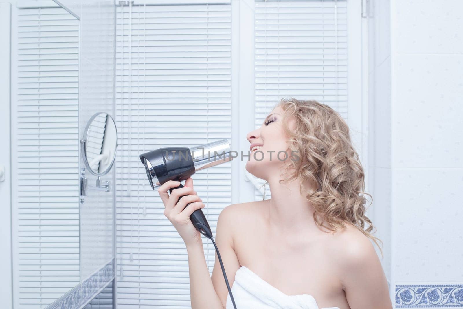 Portrait of Happy young blonde woman singing in hairdryer in bathroom