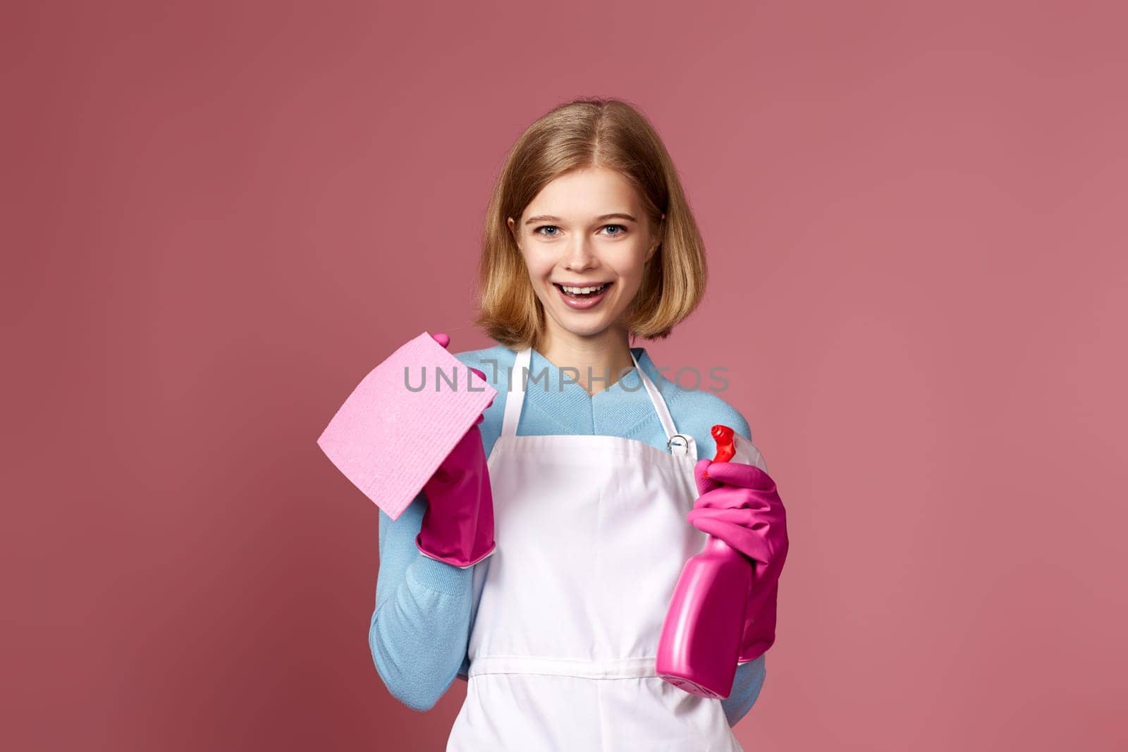 smiling blonde woman in rubber gloves with cleaning rag and detergent sprayer on pink background.
