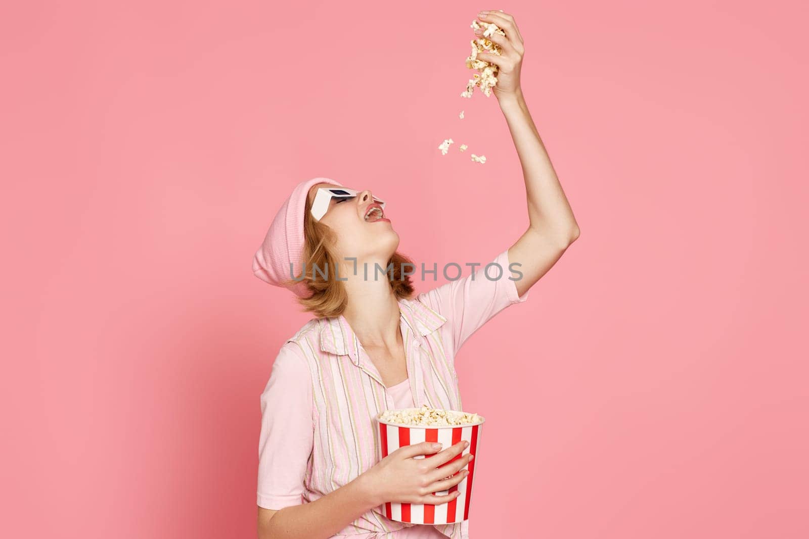 pleased blonde woman in t-shirt and hat eating popcorn on pink background.