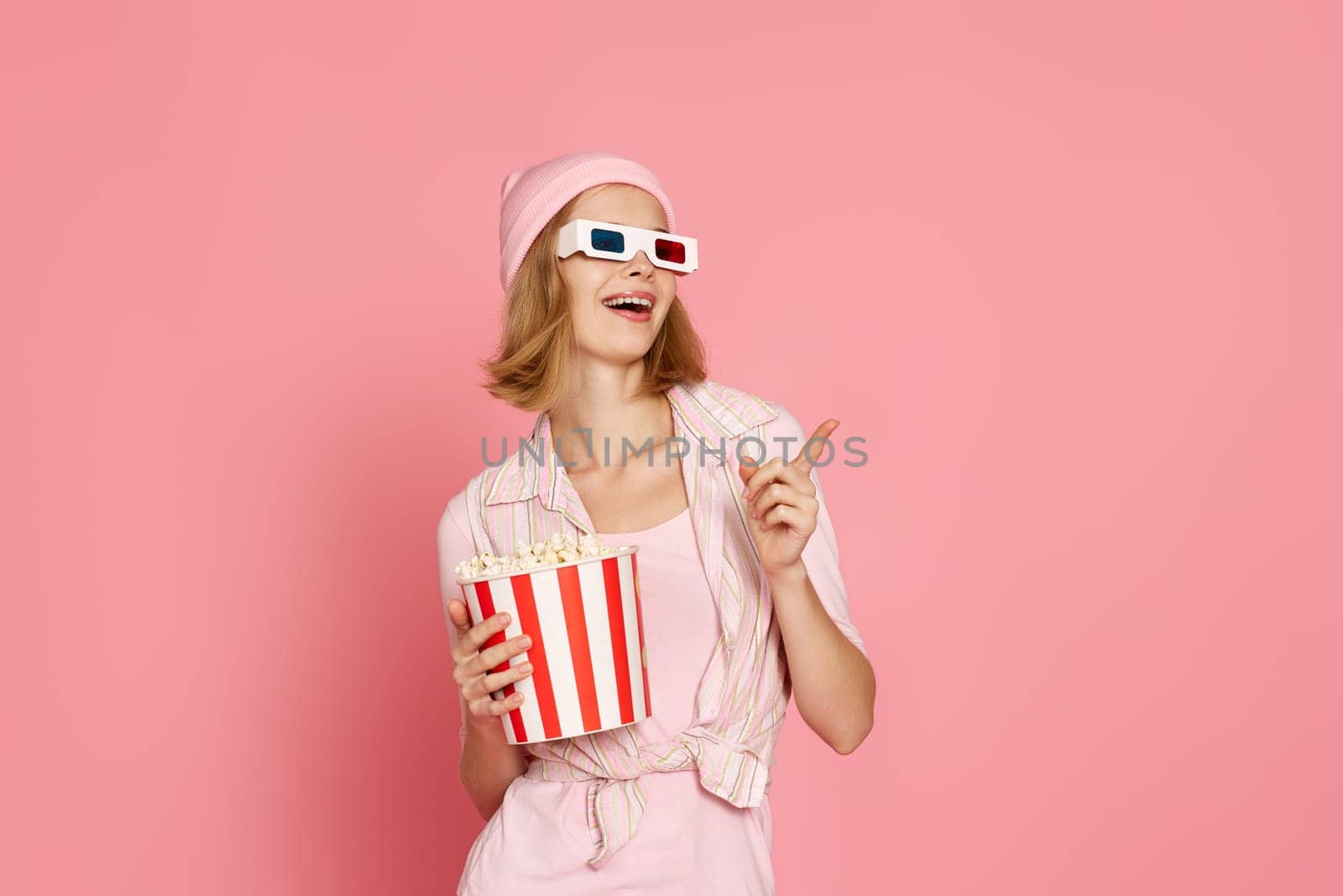 smiling woman in t-shirt and hat with popcorn pointing at the copy space on pink background.