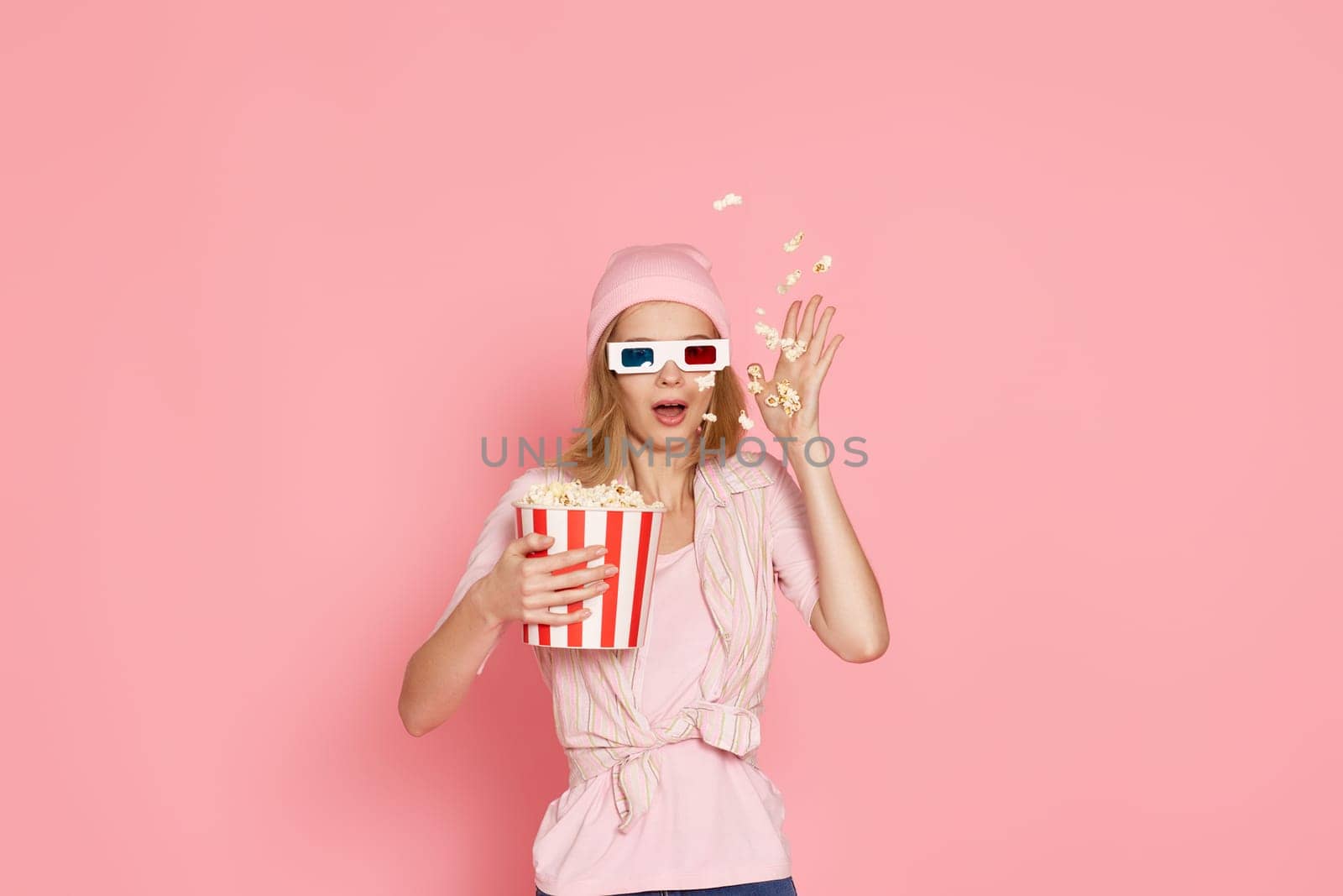terrified girl with short haircut watching horror movie , holding bucket of popcorn on pink background