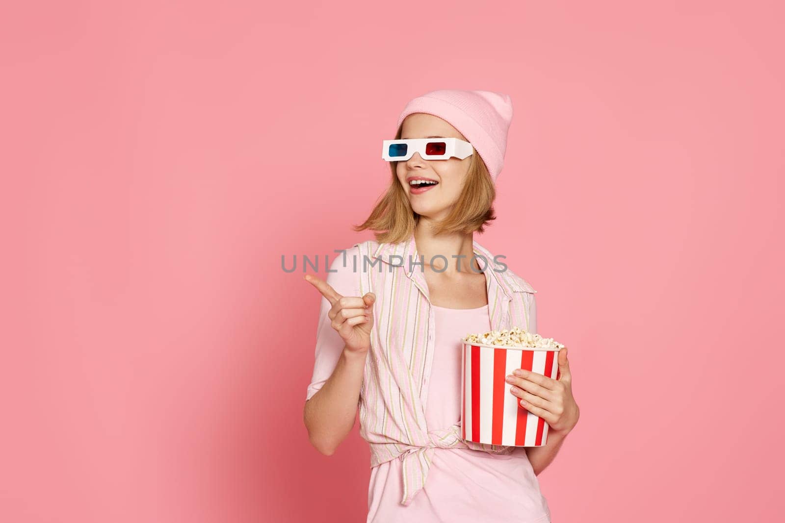 smiling woman in t-shirt and hat with popcorn pointing at the copy space on pink background.