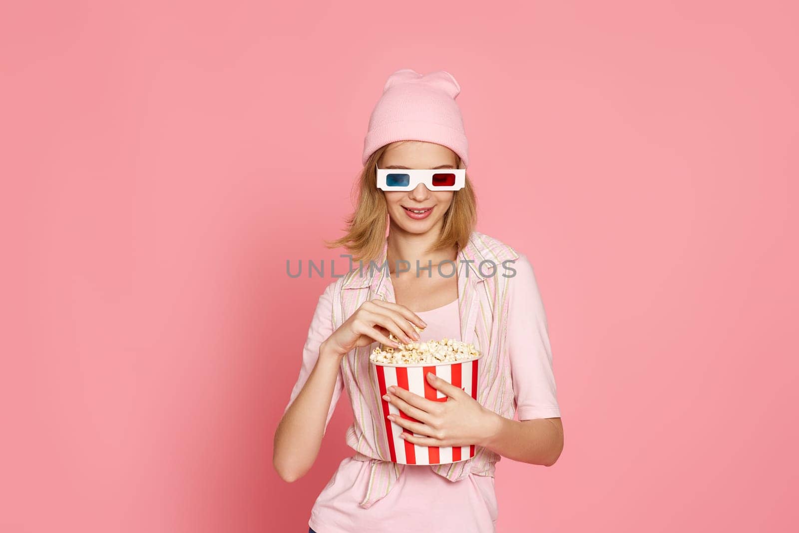 attractive blonde woman in 3d glasses holding bucket of popcorn isolated on pink background