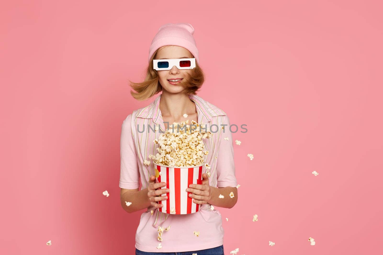 Cheerful blonde woman in 3d-glasses holding bucket of popcorn and jumping on pastel pink background.