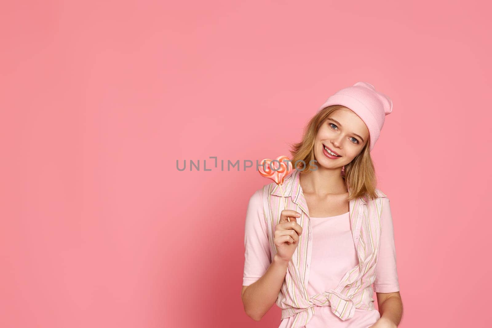 beautiful blonde woman holding big heart lollipop on pink background. copy space
