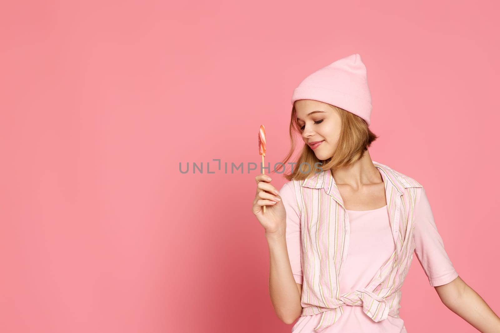 beautiful blonde woman holding colorful caramel candy on stick on pink background. copy space