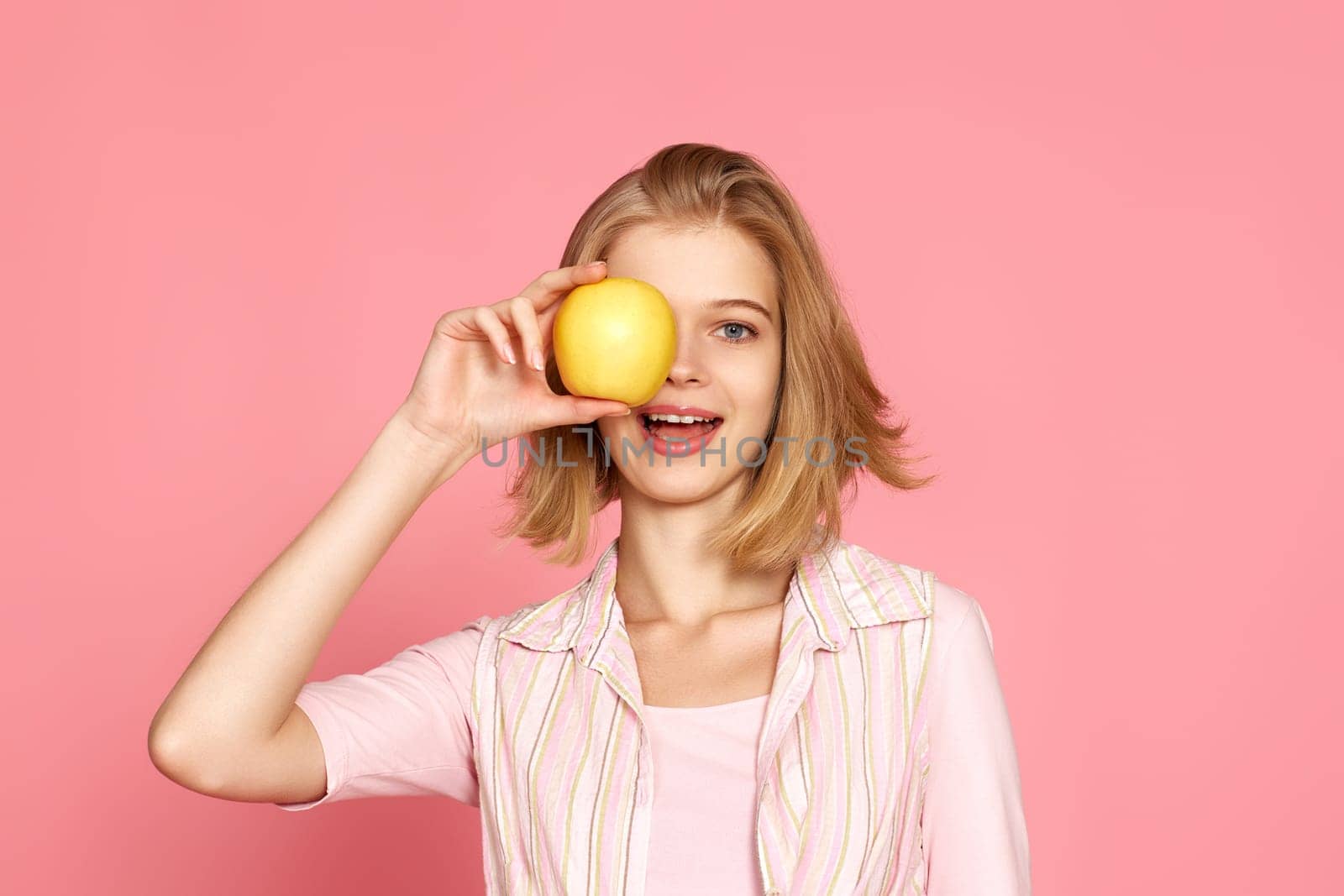 Portrait of smiling cheerful blonde woman covers one eye with fresh apple on pink background.