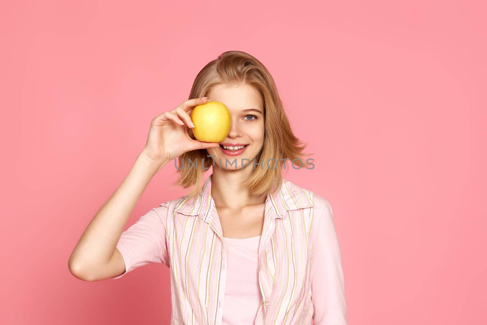 Portrait of smiling cheerful blonde woman covers one eye with fresh apple on pink background.