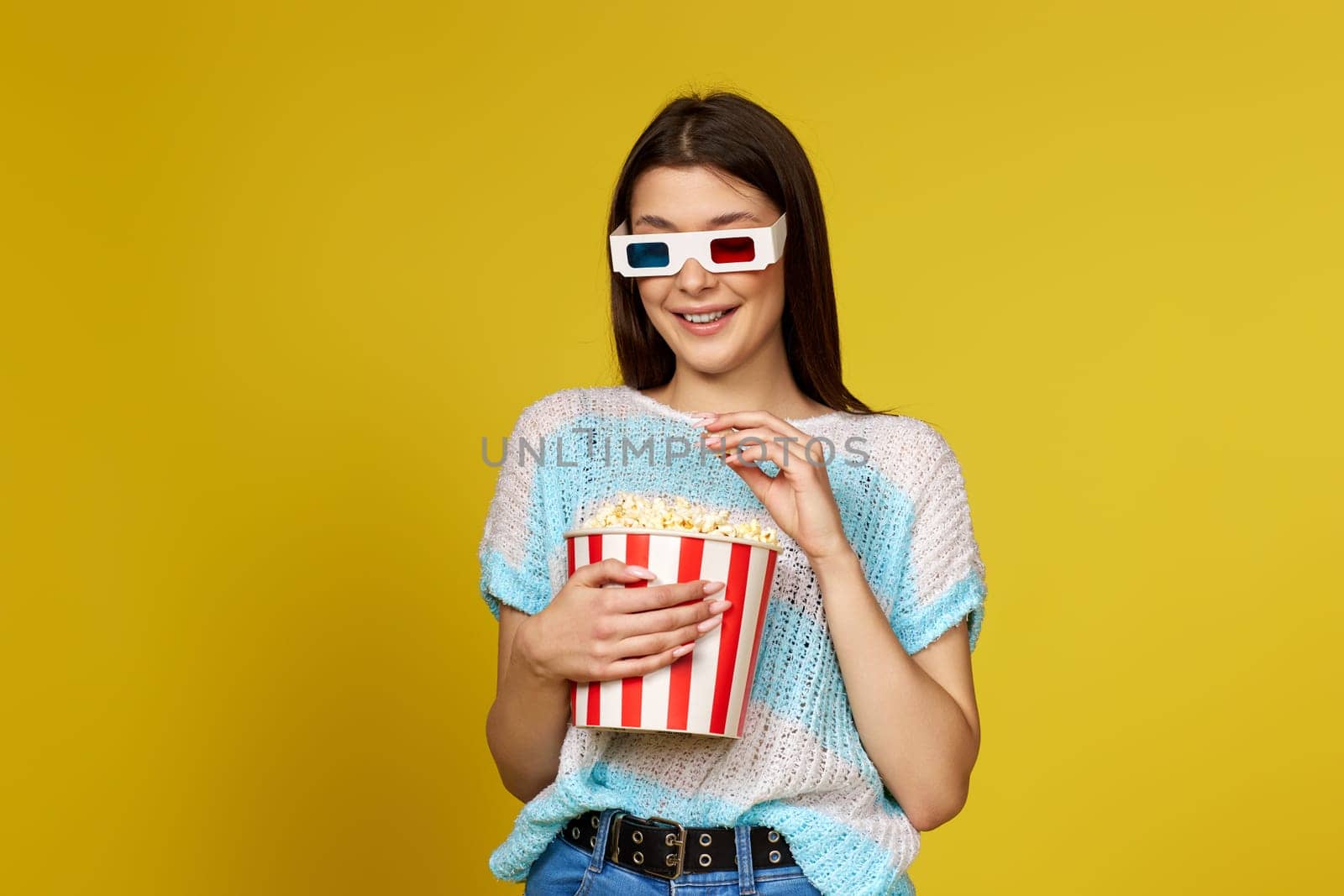 beautiful brunette girl in blue pullover eating popcorn isolated on yellow background