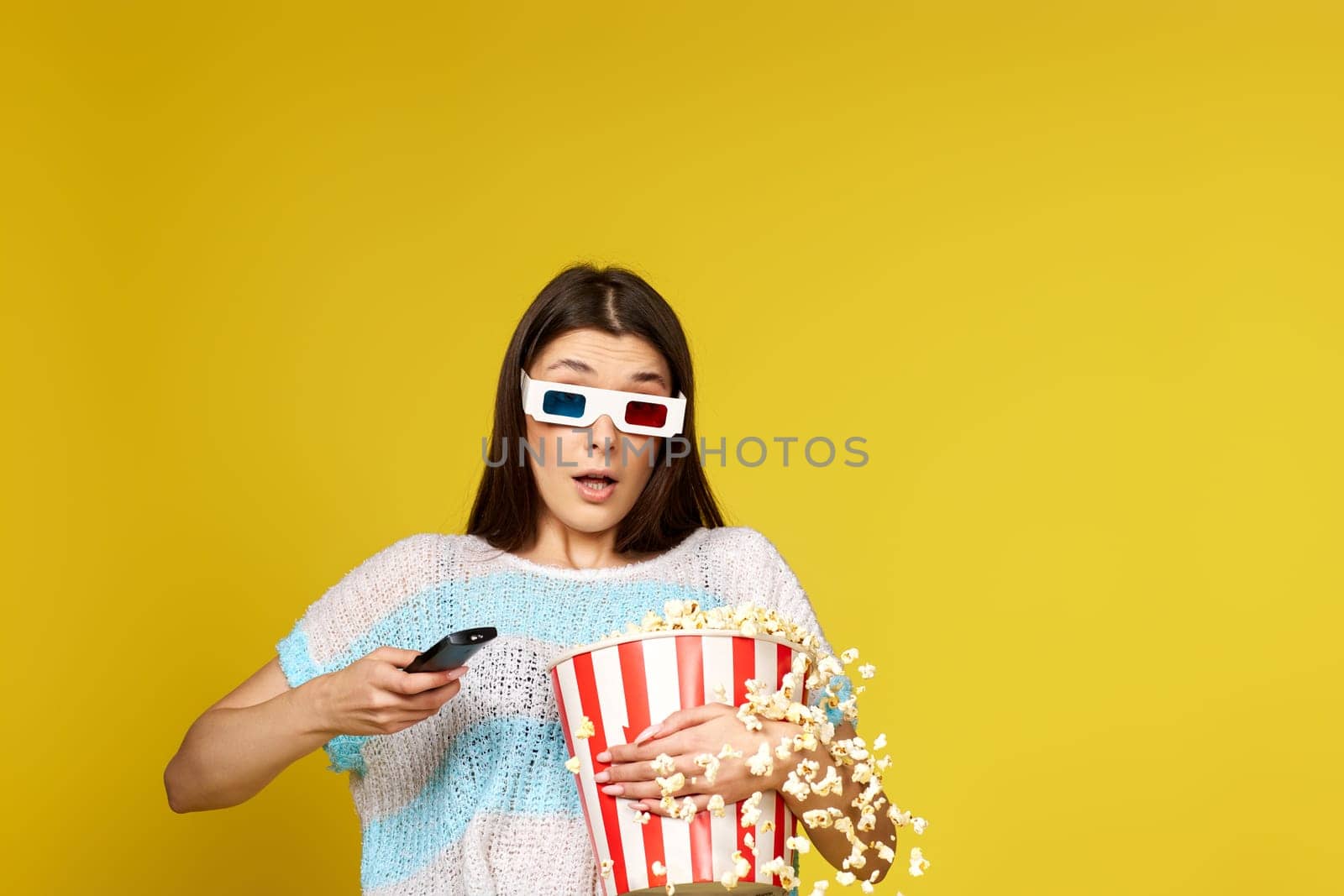 pretty woman with remote control holding bucket of popcorn and jumping on yellow background.