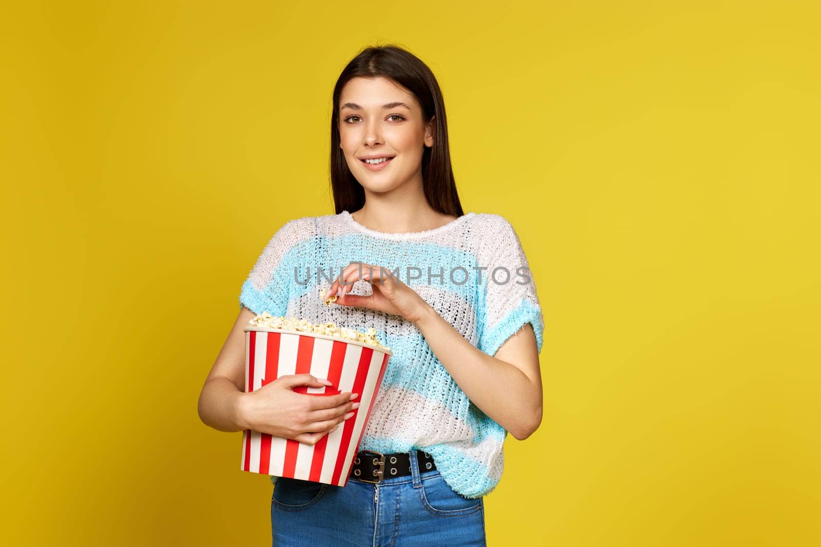 beautiful brunette girl in blue pullover eating popcorn isolated on yellow background
