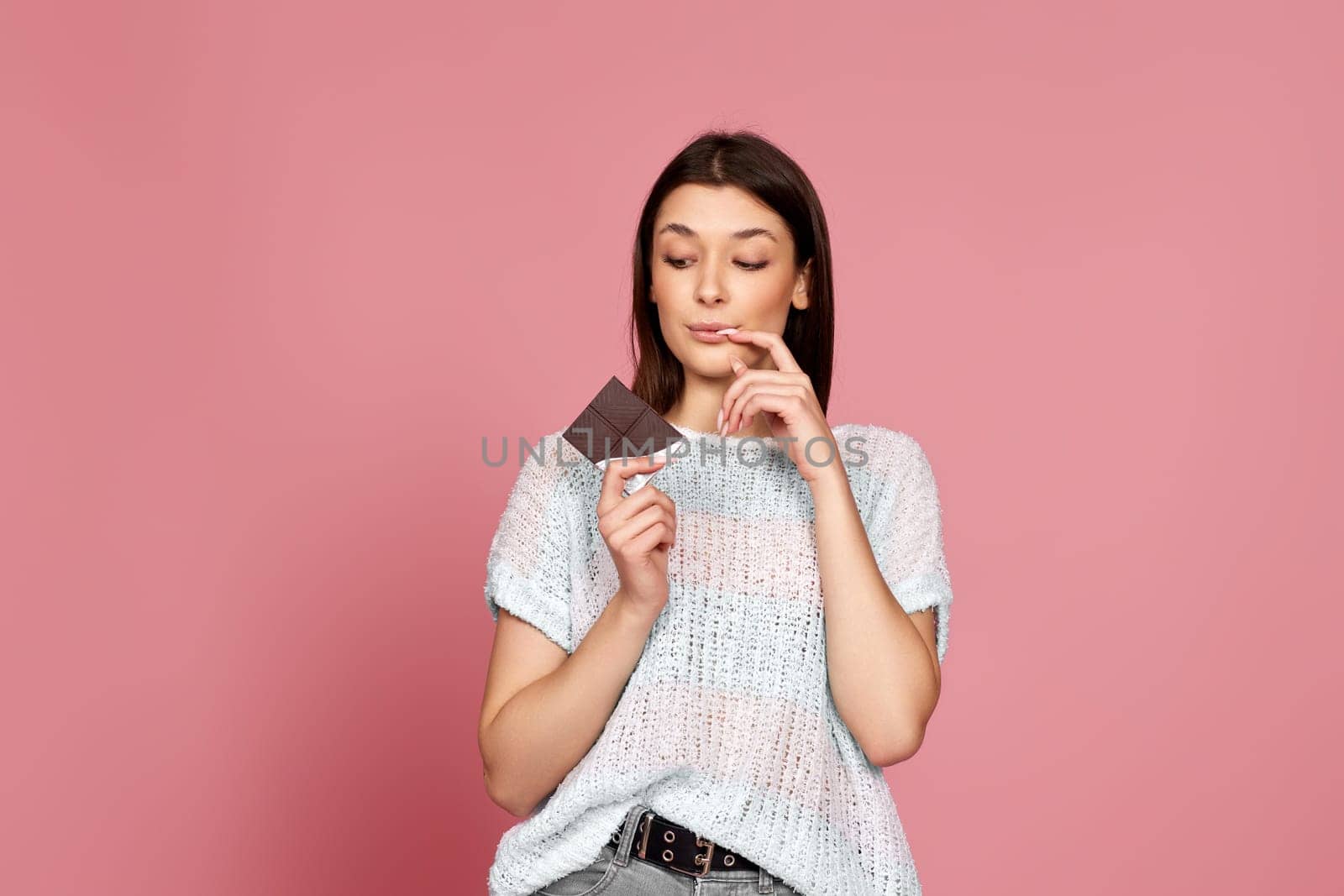 young woman in sweater holding delicious chocolate bar on pastel pink background