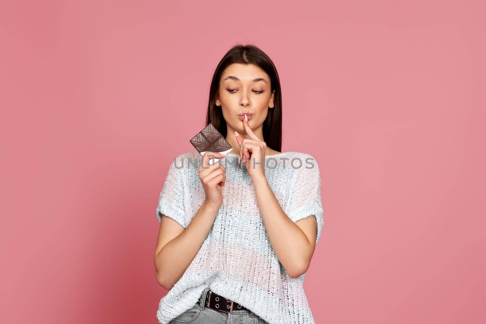 young woman in sweater holding delicious chocolate bar on pastel pink background