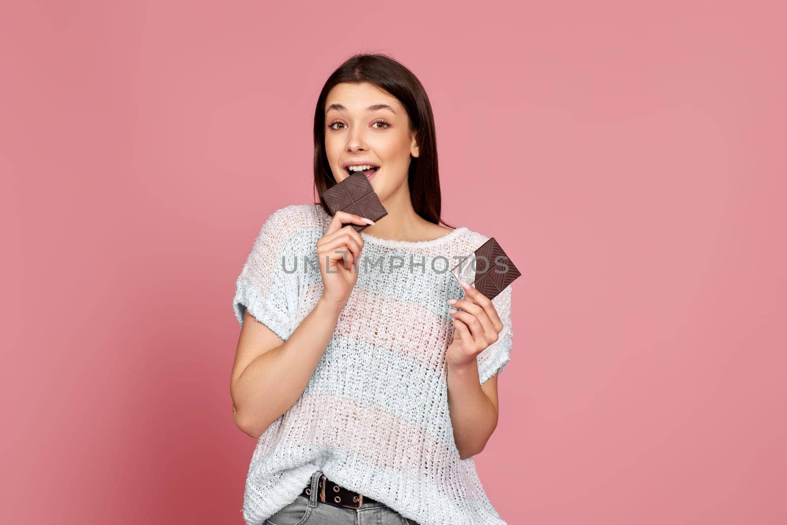 caucasian woman in sweater biting yummy chocolate bar and looking at camera on pastel pink background