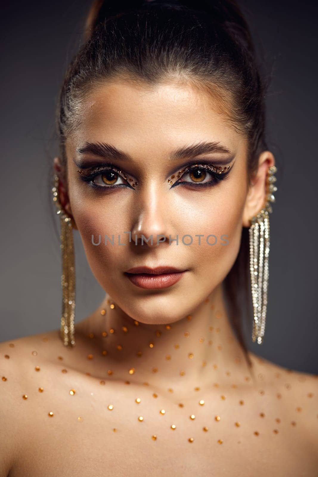 elegant fashionable model woman with creative eye makeup with arrows and rhinestones in long earring looking at the camera on gray background