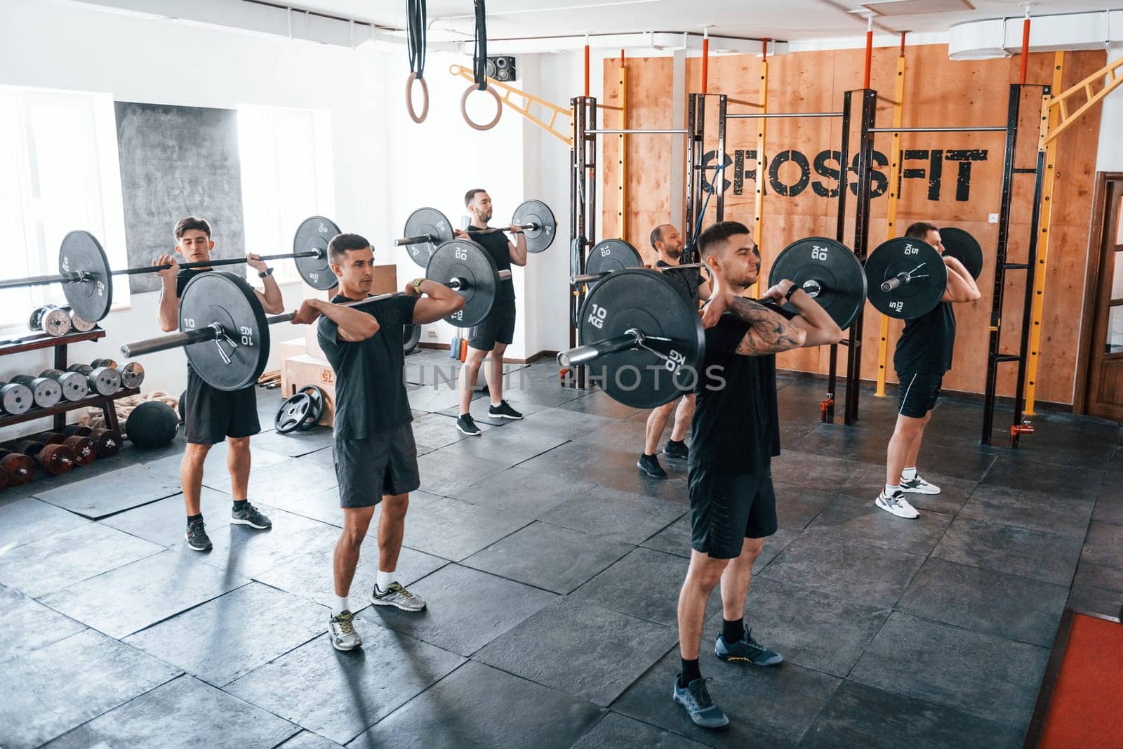Together with barbells. Group of young sportive people have crossfit day indoors in gym.