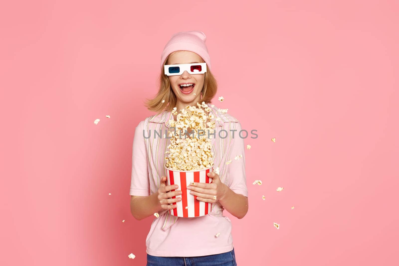 Cheerful blonde woman in 3d-glasses holding bucket of popcorn and jumping on pastel pink background.