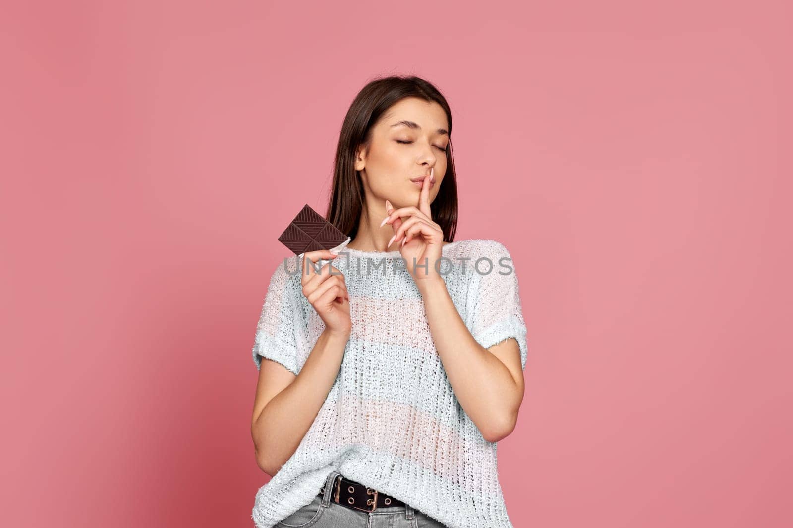 young woman in sweater holding delicious chocolate bar shows a gesture quietly on pastel pink background