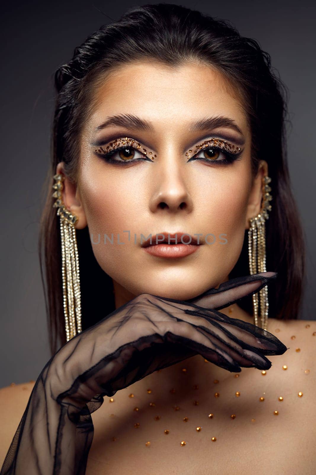 elegant fashionable model woman with creative eye makeup with arrows and rhinestones in long earring looking at the camera on gray background
