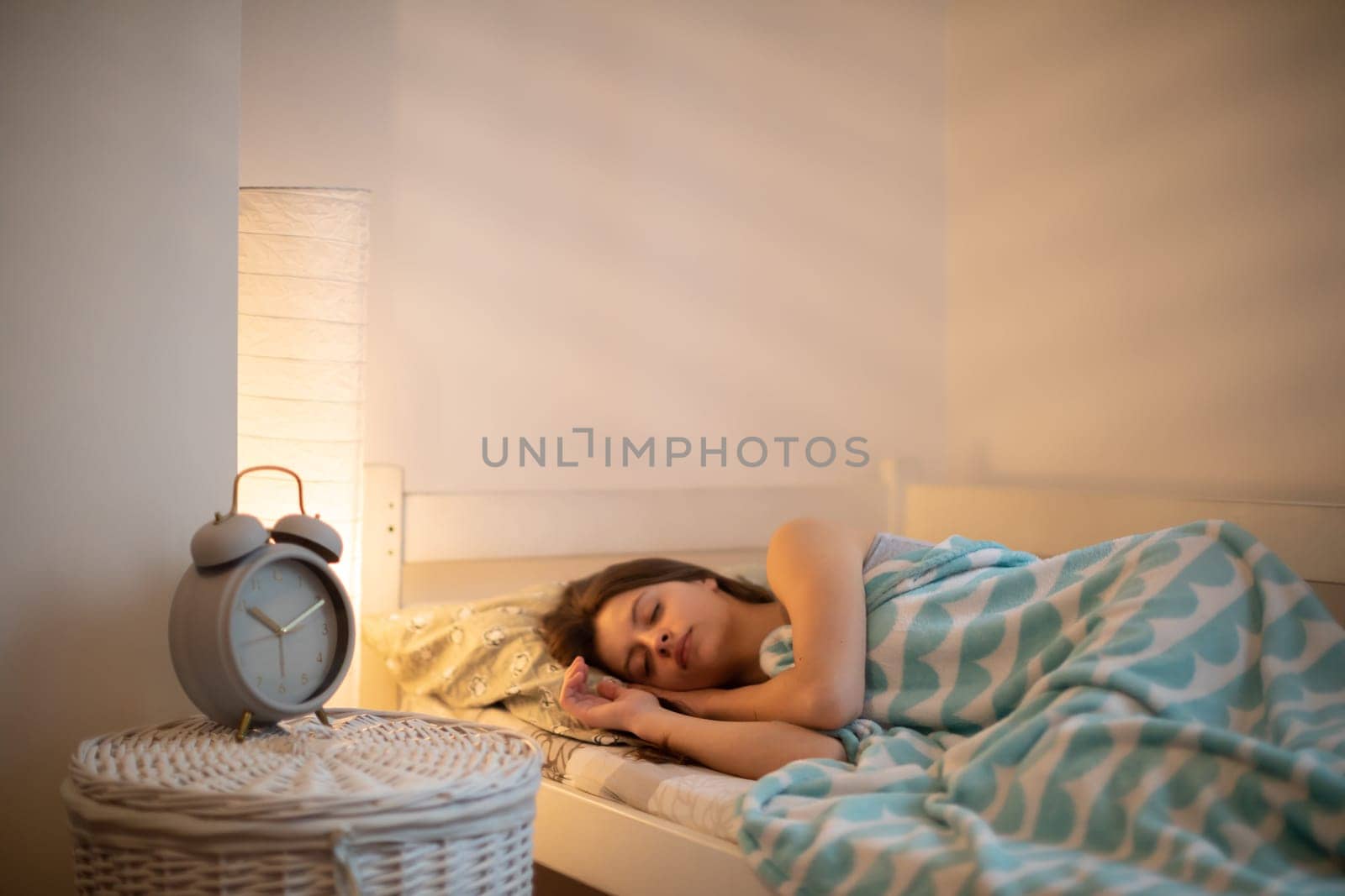 The woman sleeps in her bed with the bedside lamp lit. A young woman is lying on a bed in her bedroom, her eyes closed. A large alarm clock stands on the bedside table.